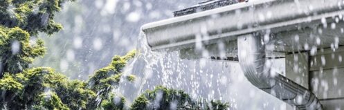 Drenching downpour rain storm water is overflowing off the tile shingle roof - streaming, rushing and splashing out over the overhanging eaves trough aluminum roof gutter system.